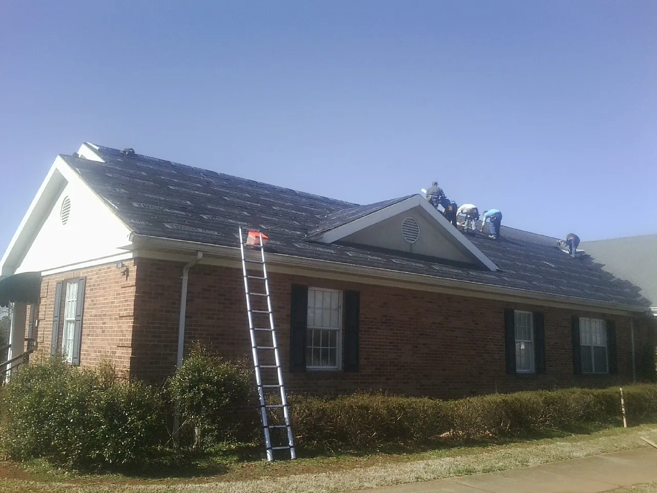 Crew tearing shingles off a brick ranch roof with a ladder set — Emergency Roof Repair work by our crew in Plum Branch