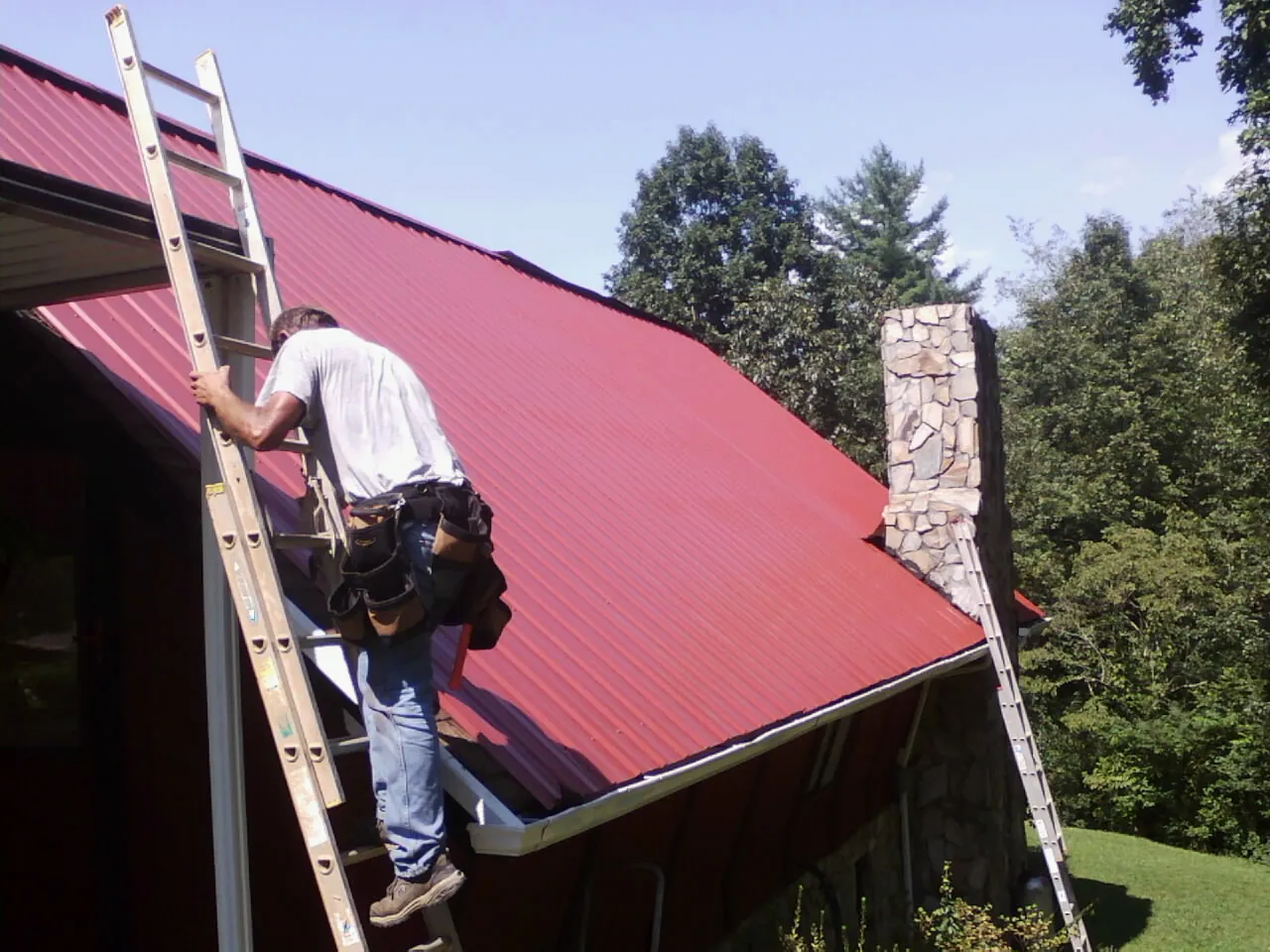Roofer on a ladder beside a red metal roof and stone chimney — Soffit & Fascia Repair by Roofing Brothers in Plum Branch